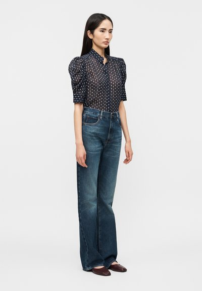 Γυναίκα που φοράει μια διάφανη μπλε μπλούζα με λευκά μοτίβα αστεριών, μανίκια με φουσκοι, ψηλόμεσο ευρύ και μακρύ τζιν μπλε, και καφέ παπούτσια, στέκεται ακίνητη.