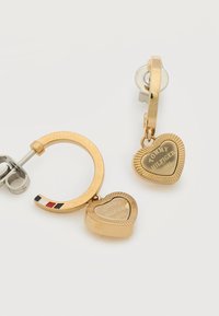 Κρεμαστά σκουλαρίκια κρίκοι από χρυσό με καρδιές-μενταγιόν χαραγμένα με "Tommy Hilfiger 1985" και μπλε, λευκή, κόκκινη ρίγα στην εξωτερική άκρη ενός κρίκου.