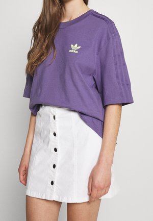 Vrouw draagt een paarse Adidas T-shirt met drie strepen op de mouwen en een witte minirok met knopen aan de voorkant.