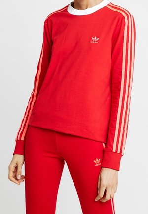 Personne portant un survêtement rouge Adidas avec trois bandes roses sur les manches et le pantalon, logo Adidas sur la poitrine et la cuisse.