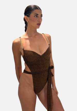 Donna con capelli intrecciati che indossa un costume intero marrone con spalline a catena dorata e un taglio laterale annodato in vita.