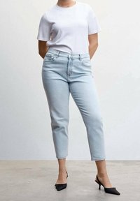 Personne portant un jean taille haute bleu clair, un t-shirt blanc à manches courtes et des escarpins noirs à bouts pointus, debout devant un fond blanc.