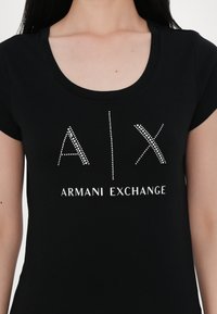 T-shirt noir à manches courtes avec un col rond et un logo scintillant "A|X ARMANI EXCHANGE" en clous argentés sur le devant.