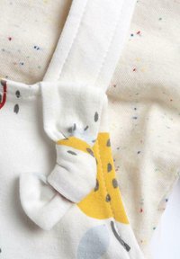 Tela blanca con motas multicolores; presenta un detalle de correa redonda con un patrón amarillo y gris, textura suave y secciones fruncidas.