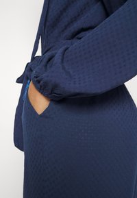 Navy blue garment with textured fabric, featuring a relaxed fit, elastic cuffs, and a side pocket detail. Consistent pattern throughout.