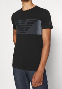 Svart T-shirt i bomull med ett stort grått emblem på bröstet och textdetaljer på ärmen. Kort ärm och rundad halsringning.