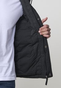 Melnā pūkainā jaka iekšpuse, kurā redzama izšūta tekstūra, rāvējslēdzēja aizdare, pogas ar spiedpogu un iekšēja kabata kreisajā pusē.