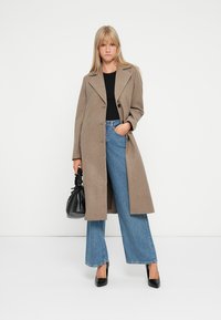 Manteau en laine marron avec un col en pointe et des boutons à l'avant, associé à un jean en denim bleu taille haute et à pattes d'éléphant, ainsi qu'à des talons noirs à bout pointu.