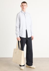 Lys blåstripet skjorte, svarte vide bukser, beige handlenett med to håndtak, og nøytrale sko på en nøytral bakgrunn.