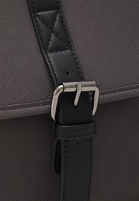 Borsa di tessuto grigia con una tracolla in pelle nera, caratterizzata da una fibbia in metallo argentato. La tracolla include un accento testurizzato e cuciture precise.