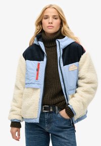 Giacca puffer azzurro chiaro con dettagli in felpa blu navy e crema, dotata di tasca con zipper, polsini a coste e patch con logo montagna. Maglione marrone sotto.