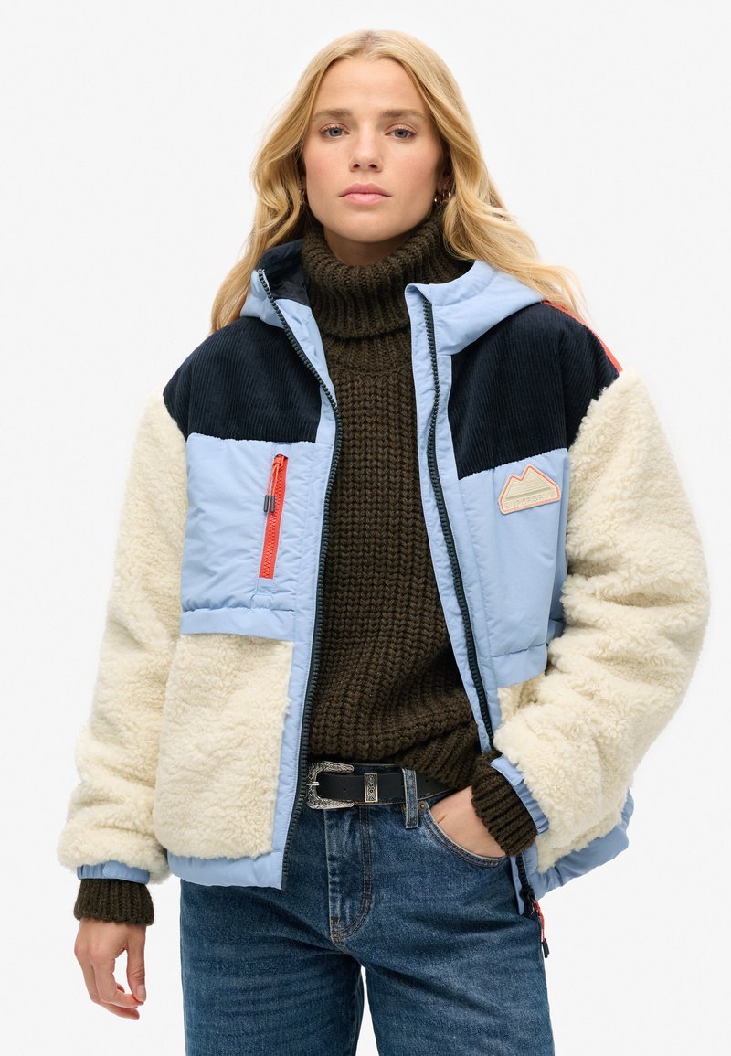 Giacca puffer azzurro chiaro con dettagli in felpa blu navy e crema, dotata di tasca con zipper, polsini a coste e patch con logo montagna. Maglione marrone sotto.