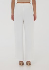 Pantaloni bianchi a gamba larga con una finitura testurizzata, una vita elastica e un fit rilassato; indossati con tacchi neutri, mostrando il dettaglio della caviglia.