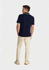 Young man stands with his back to the camera, wearing a navy polo shirt and light beige pants. He has short hair and sport shoes.