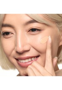 Femme souriante aux cheveux clairs applique un fond de teint liquide beige sur sa joue droite avec l'index.