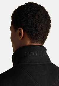 Giacca in fleece nera con colletto alto e branding "JOULES" in rilievo, tessuto testurizzato e dettagli di cucitura visibili.