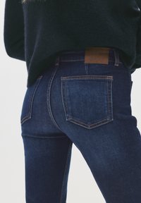 Sötétkék farmernadrágot és fekete pulóvert viselő személy, hátulról mutatva, deréktól a comb közepéig terjedő nézetben, hosszan növesztett hajjal.