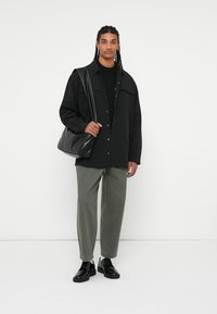 Černá oversized košile na knoflíky, černý pletený svetr pod ním, zelené tapered kalhoty, černá kožená kabelka přes rameno, lesklé černé boty.
