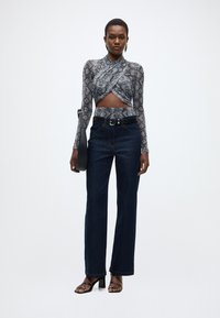 Crop top gris à manches longues imprimé serpent avec un design noué, associé à un jean denim bleu foncé taille haute et des sandales noires à talons blocs.