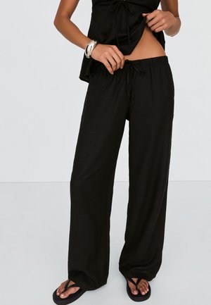 Femme portant un pantalon ample noir à cordon, un haut noir, un bracelet argenté et des tongs noires, ajustant son pantalon à la taille.