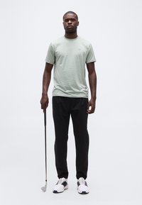 Νέος άνδρας όρθιος, κρατώντας ένα γκολφ ρόπαλο στο δεξί χέρι, φοράει ανοιχτό πράσινο μπλουζάκι, μαύρο παντελόνι και λευκά αθλητικά παπούτσια σε απλό φόντο.