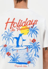 T-shirt bianco in cotone con una grafica blu e rossa di un pinguino su una tavola da surf, accompagnato da piante tropicali e un'illustrazione del sole.