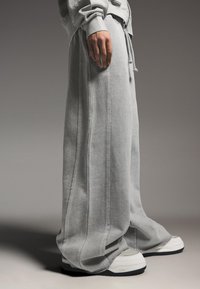 Persona che indossa pantaloni della tuta grigio chiaro oversize e sneakers bianche, in piedi davanti a uno sfondo neutro e semplice.