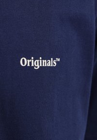 Marineblått stoff med "Originals™"-logoen i hvitt, plassert sentralt. Glatt tekstur med et fint strikkemønster.