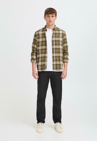 Green and beige plaid shirt over a white t-shirt, paired with black pants and light sneakers. Model stands against a plain background.