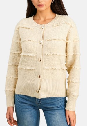 Femme portant un cardigan en maille texturée crème avec des rayures à franges décoratives et des boutons dorés, associé à un jean bleu.