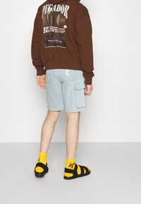 Hnědá mikina se grafickým potiskem, světle modré cargo šortky, černé sandály a žluté ponožky. Mezi významné detaily patří boční kapsy šortků.