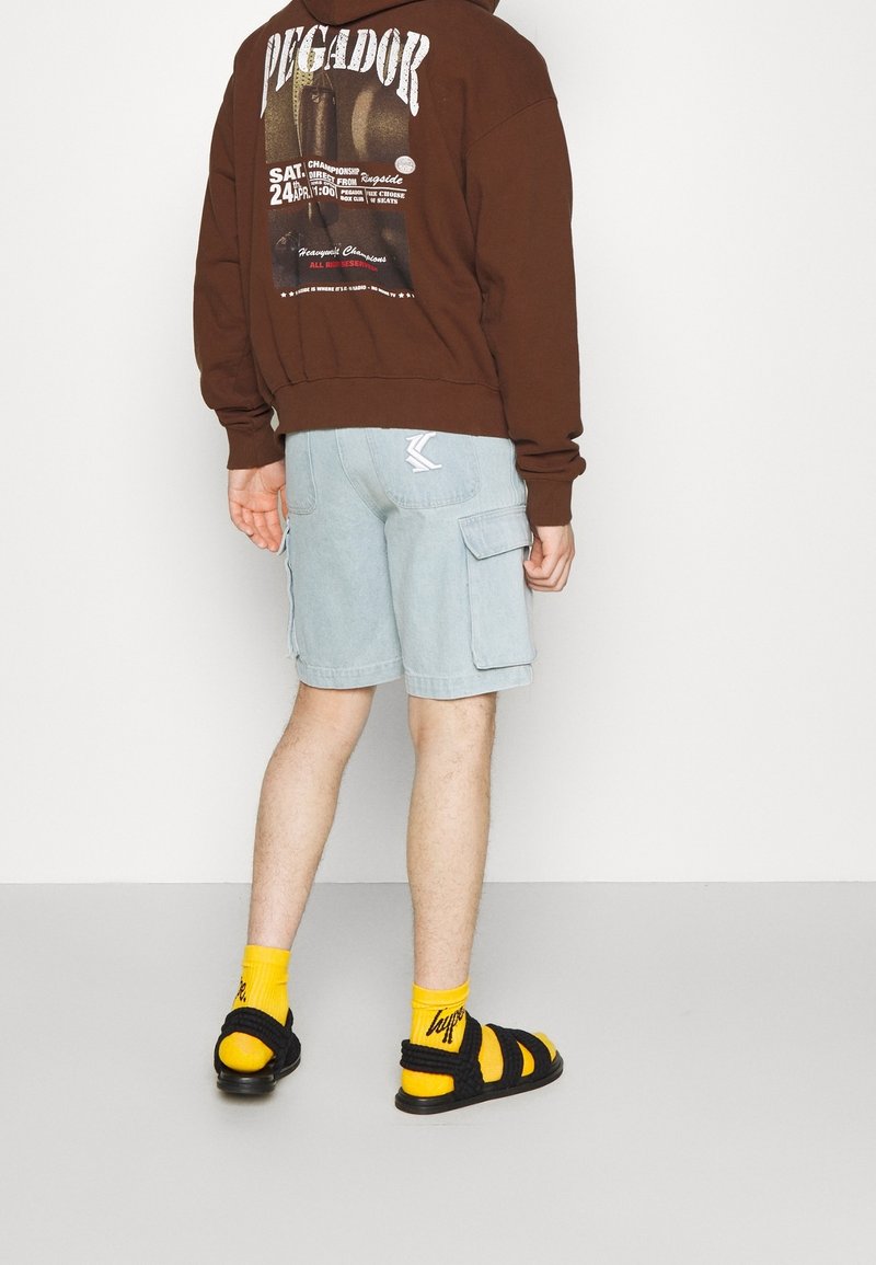 Hnědá mikina se grafickým potiskem, světle modré cargo šortky, černé sandály a žluté ponožky. Mezi významné detaily patří boční kapsy šortků.