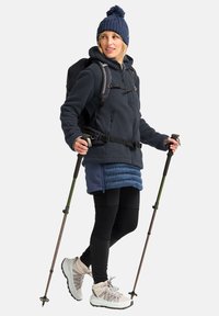 Fleecejack in donkergrijs, gelaagd met een blauwe geïsoleerde rok, zwarte leggings en lichtgekleurde wandelschoenen. Bevat trekkingstokken en een gebreide muts.