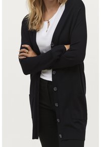 Kvinna i en svart knäppt kardigan över en vit blus och svarta byxor, med armarna korsade och en guldring på vänster hand.