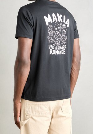 Personne portant un t-shirt noir avec des cœurs de dessin animé blancs et le texte "MAKIA Life is a Hard Romance" dans le dos, associé à un pantalon beige clair.
