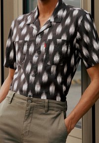 Camicia a maniche corte in grigio scuro con motivo bianco simile a piume; presenta davanti una chiusura con bottoni e due tasche sul petto, abbinata a pantaloni kaki chiari.