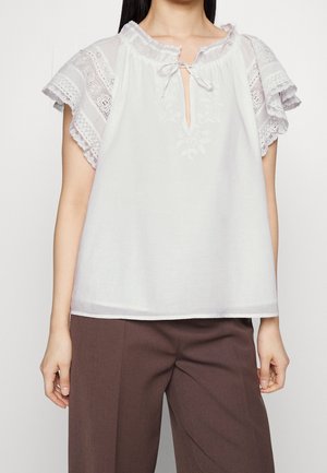 Weiße Bluse mit Spitzen-Flatterärmeln, bestickter Vorderseite, Schlitz-Ausschnitt mit Schnürung, getragen zu brauner Hose, dargestellt vom Hals bis zur Hüfte.