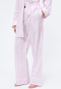 Persona vistiendo pantalones de pijama holgados a rayas rosas y blancas y camisa a juego, de pie descalza con una mano en el bolsillo.