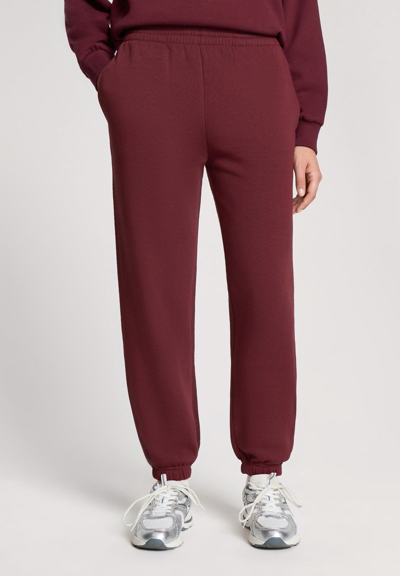 Persona che indossa pantaloni jogger marroni e sneakers bianco argento, in piedi con le mani in tasca su uno sfondo semplice.