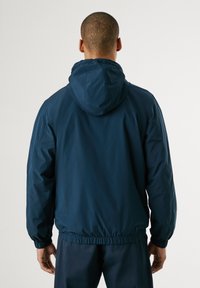 Chaqueta ligera de color azul marino con capucha, puños elásticos y cintura ajustable, que presenta una textura suave y un diseño minimalista.