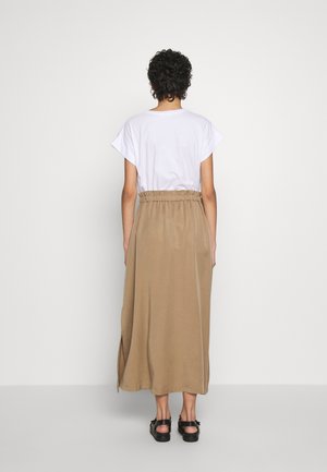 Personne aux cheveux courts et bouclés portant un haut blanc à manches courtes, une jupe midi taille haute beige et des sandales noires, de dos.