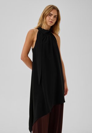 Femme aux cheveux blonds portant un haut noir asymétrique sans manches et un pantalon marron sur un fond gris clair uni.