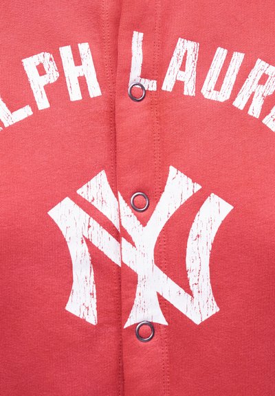 Κόκκινο ύφασμα με λευκό φθαρμένο κείμενο "Ralph Lauren" και επικαλυπτόμενο λογότυπο "NY", στο κέντρο κοντά σε τρία μεταλλικά κουμπιά τύπου snap.
