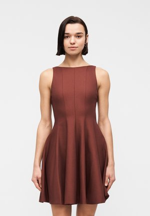 Vrouw met kort donker haar die een mouwloze, aansluitende bruine jurk draagt met verticale naaddetails en een uitlopende rok, staand tegen een effen achtergrond.