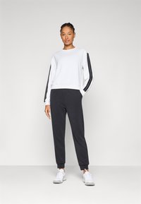 Λευκή μπλούζα με μαύρες πλευρικές ρίγες, συνδυασμένη με σκούρες γκρι φόρμες και λευκά αθλητικά παπούτσια. Μαλακό ύφασμα με άνετη εφαρμογή.