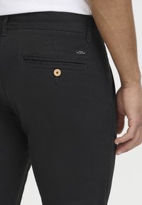 Gros plan sur la poche arrière d'un pantalon noir avec un bouton en bois et une petite étiquette portant la mention « All day Every day denim » sur le côté du bras d'une personne.