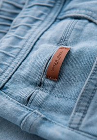 Tissu en denim bleu clair présentant une surface texturée et une couture. Une étiquette en cuir marron est attachée, affichant les détails de la marque.