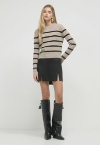 Donna in piedi con un maglione a righe beige e nero, una gonna nera con spacchi frontali e stivali neri in pelle al ginocchio con frange.