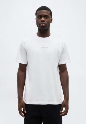 Giovane uomo con capelli corti che indossa una maglietta bianca a maniche corte semplice e pantaloni neri, in piedi davanti a uno sfondo chiaro e uniforme.