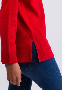 Der rote gestrickte Pullover hat gerippte Bündchen und einen gerippten Saum, mit seitlichen Schlitzen für mehr Bewegungsfreiheit. Kombiniert mit dunkelblauen, figurbetonten Jeans, die eine glatte Textur zeigen.